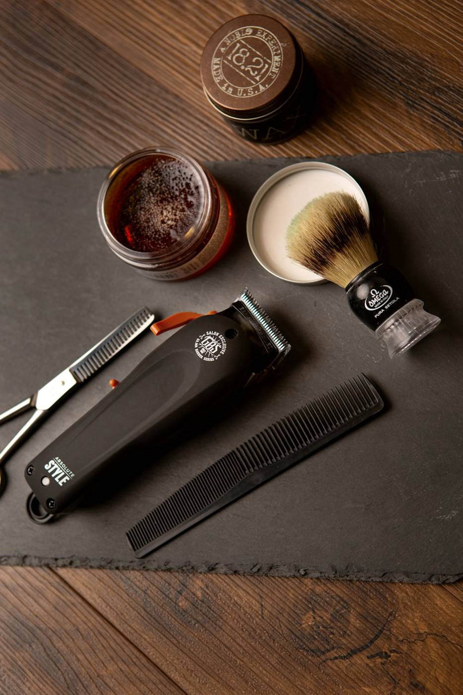 Pomade jar, shaving brush and clippers on a dark board