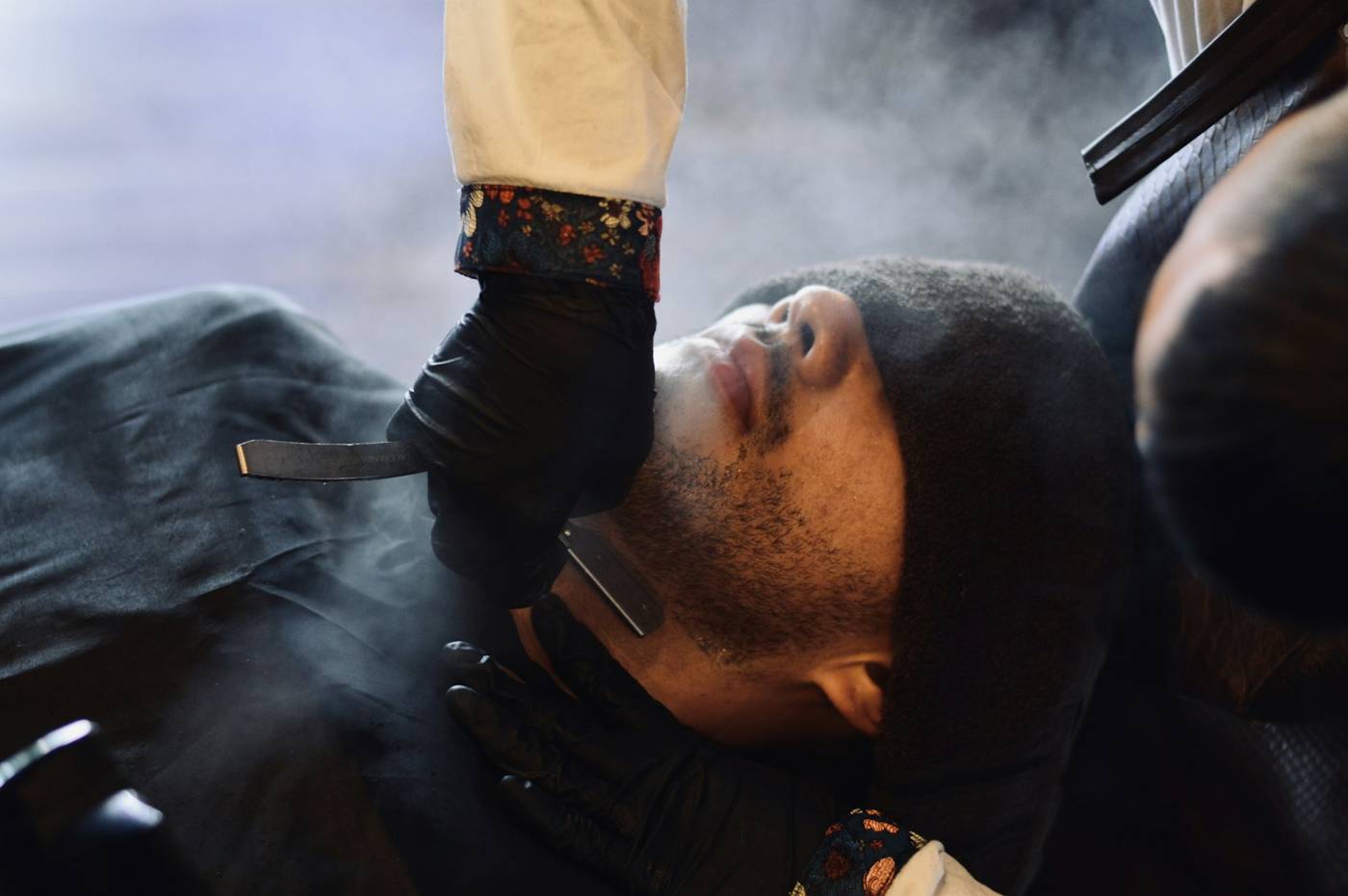 Barber shaving a client with a straight razor as steam rises from a hot towel