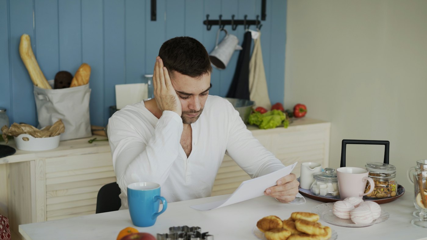 A man at his kitchen table reading a letter with a worried look