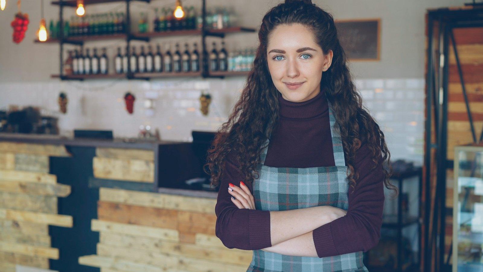 A smiling cafe owner in an apron standing with arms crossed behind her counter