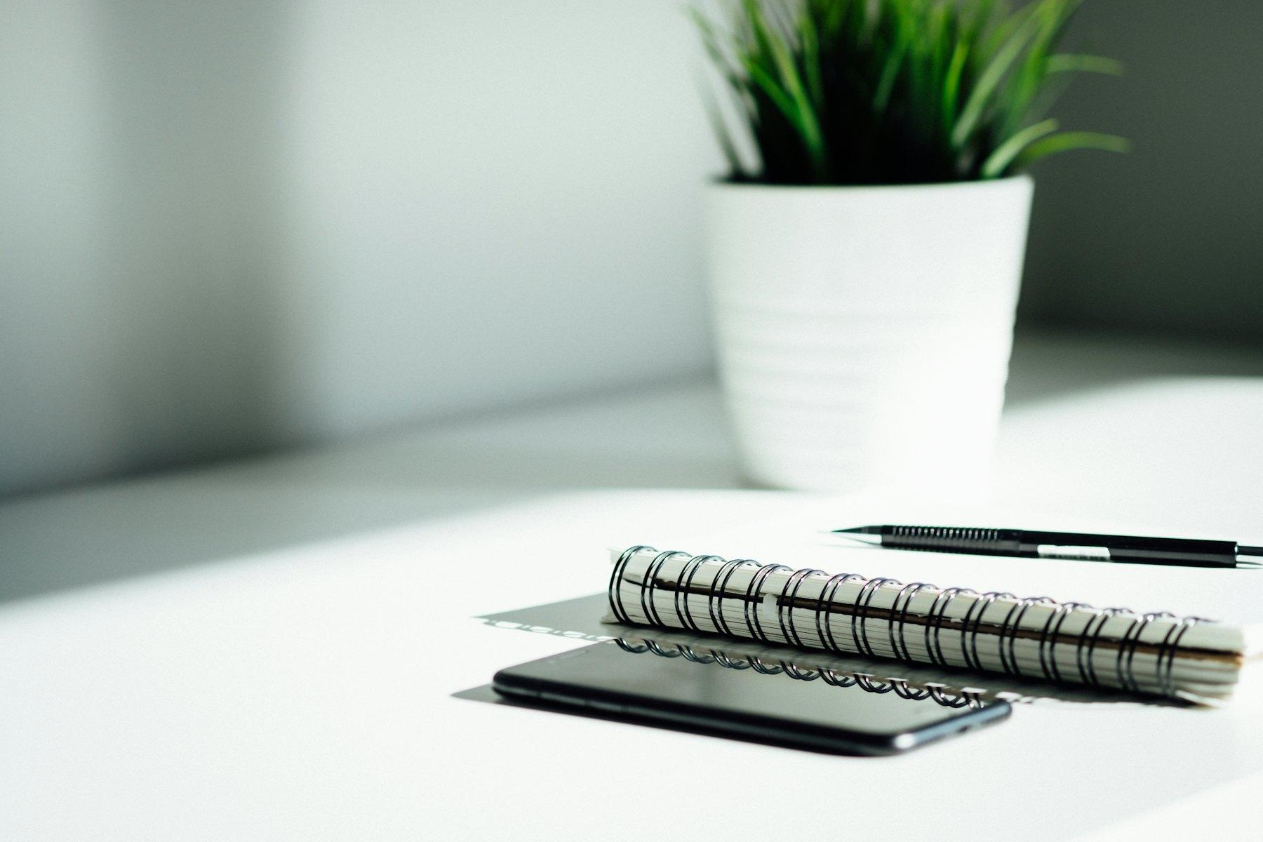 A spiral notebook, pen and phone on a white desk beside a potted plant in soft daylight