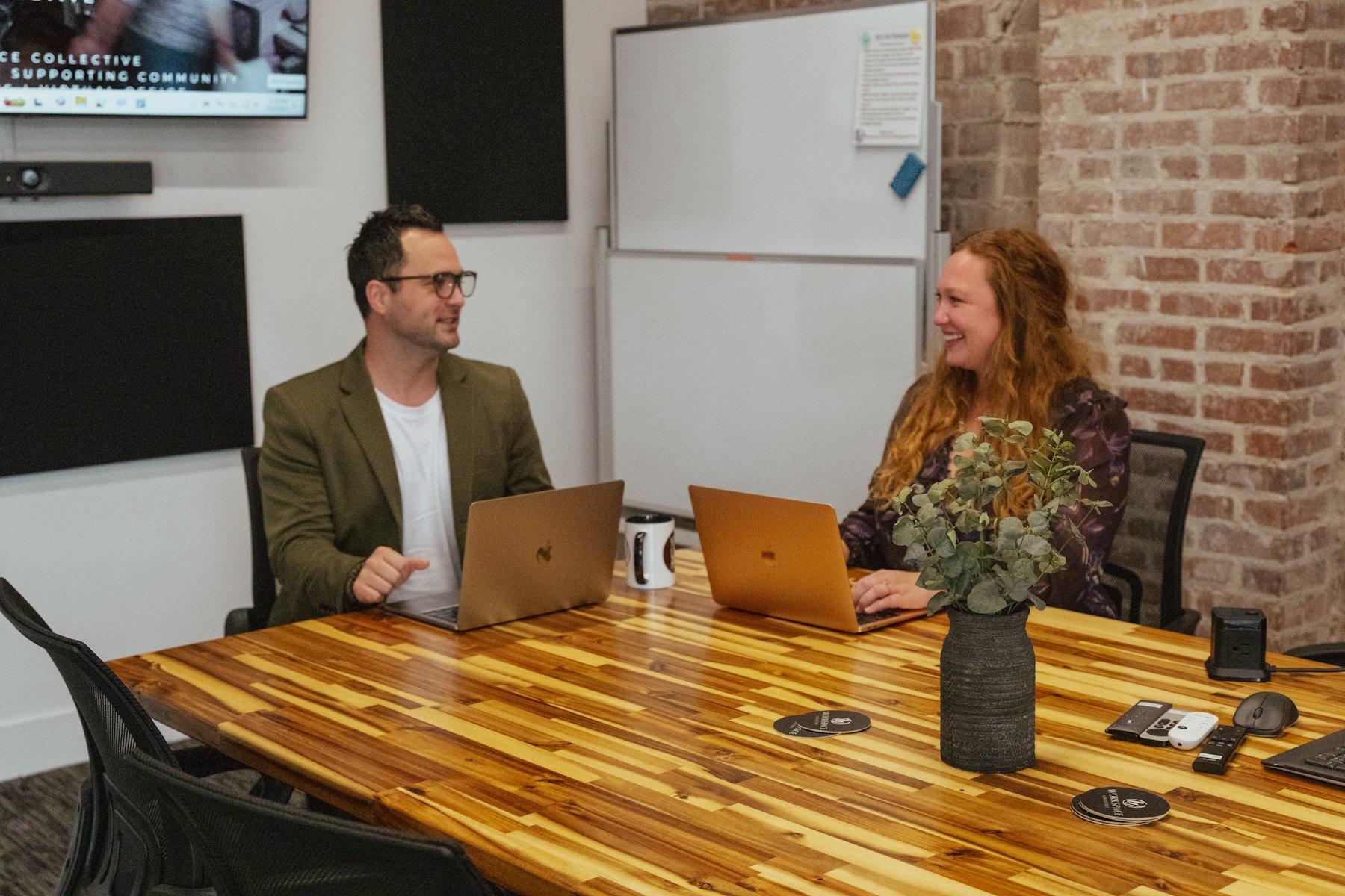 Two people talking and smiling at a wooden meeting table with laptops open