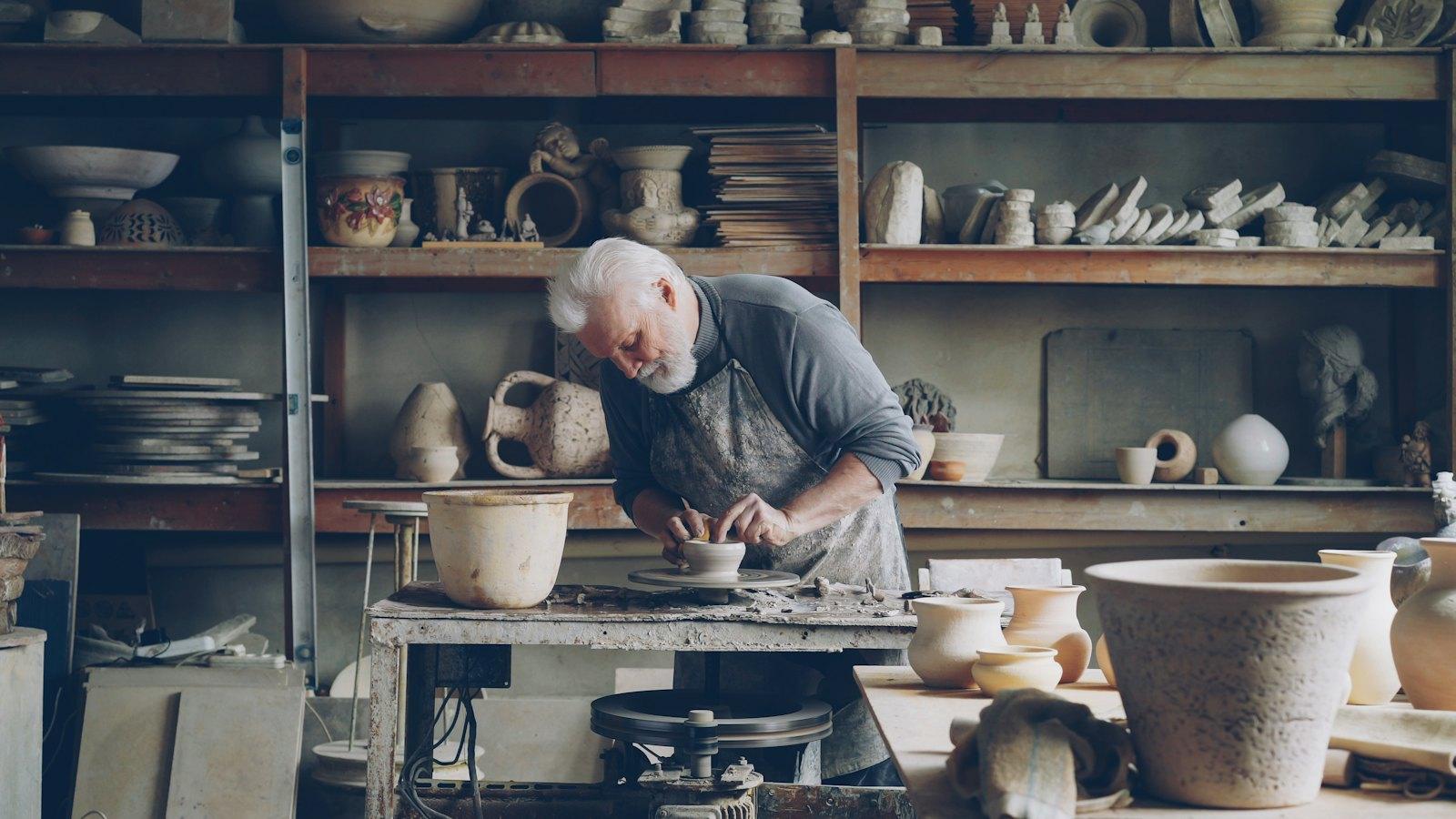 A ceramicist shaping clay at a wheel in a workshop lined with shelves of pots