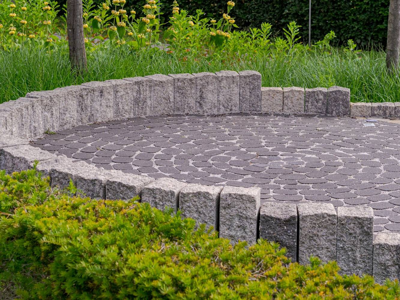 A finished round stone patio edged with a low granite wall and green shrubs