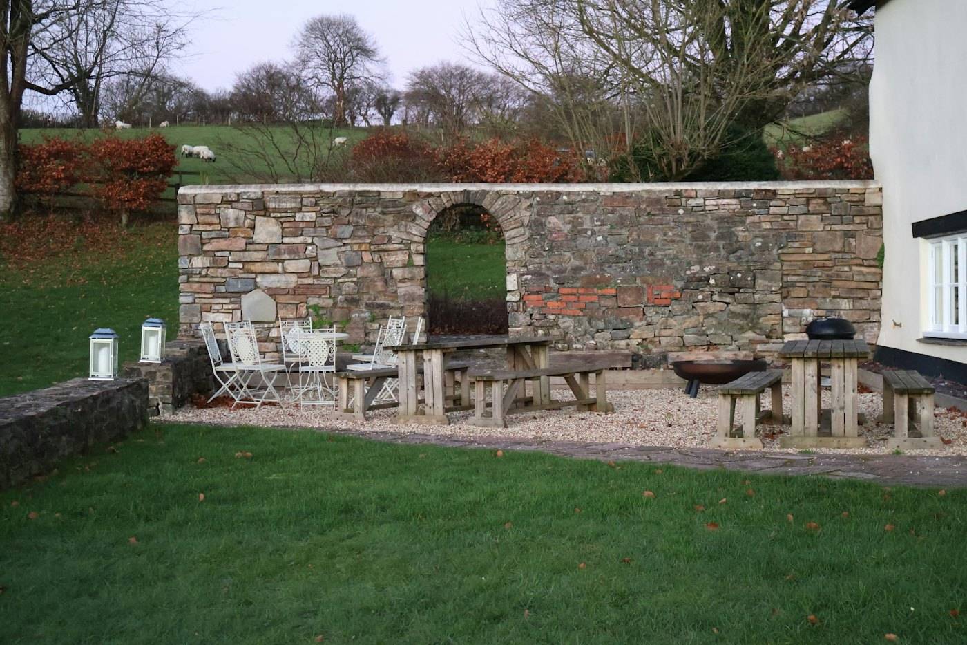 A dry stone wall with an arched opening behind a gravel courtyard with tables and chairs