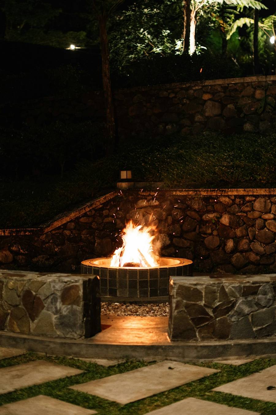 A round stone fire pit glowing at night inside a low stone wall