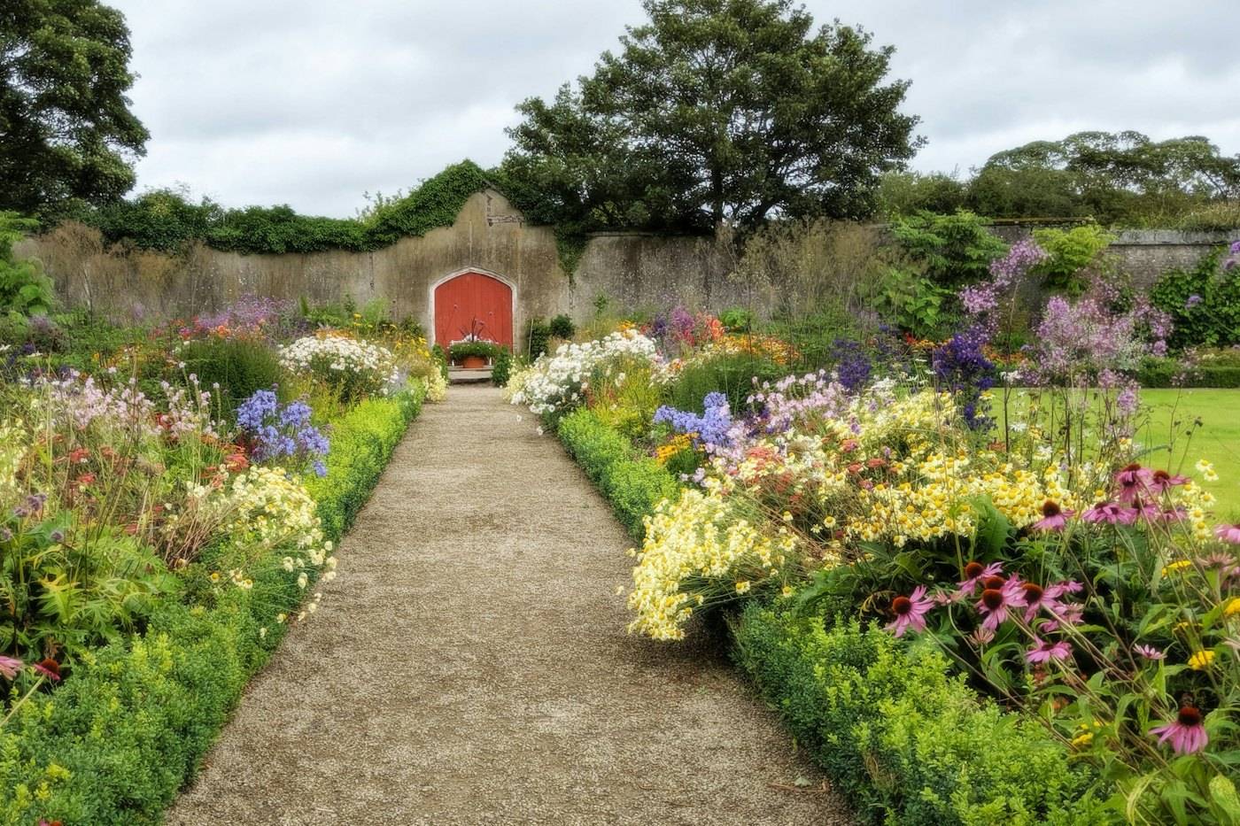 A straight path between two deep flower borders leading to a red garden door