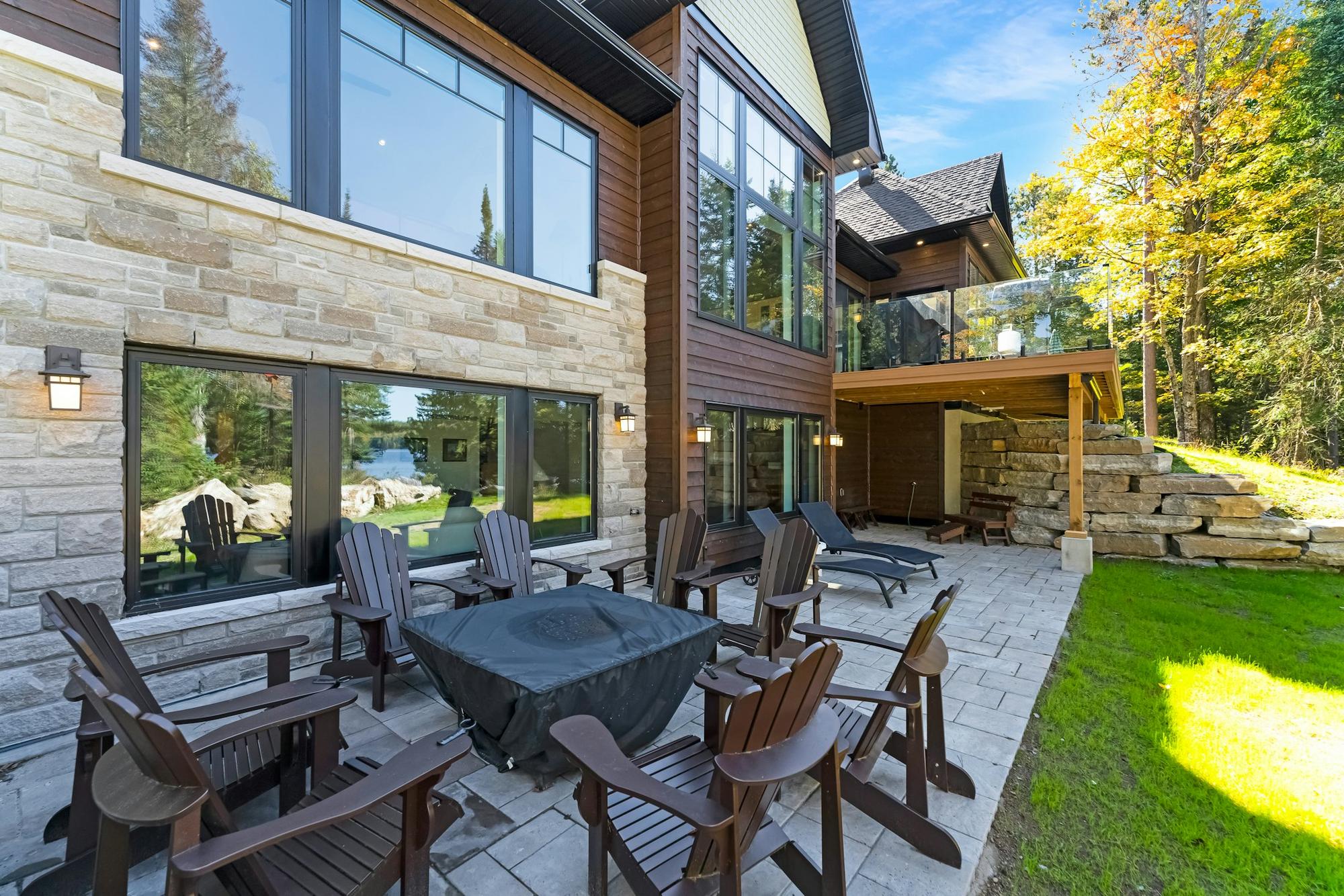 A stone patio with dark wooden chairs and a fire pit behind a stone and timber house, with lawn and trees beyond