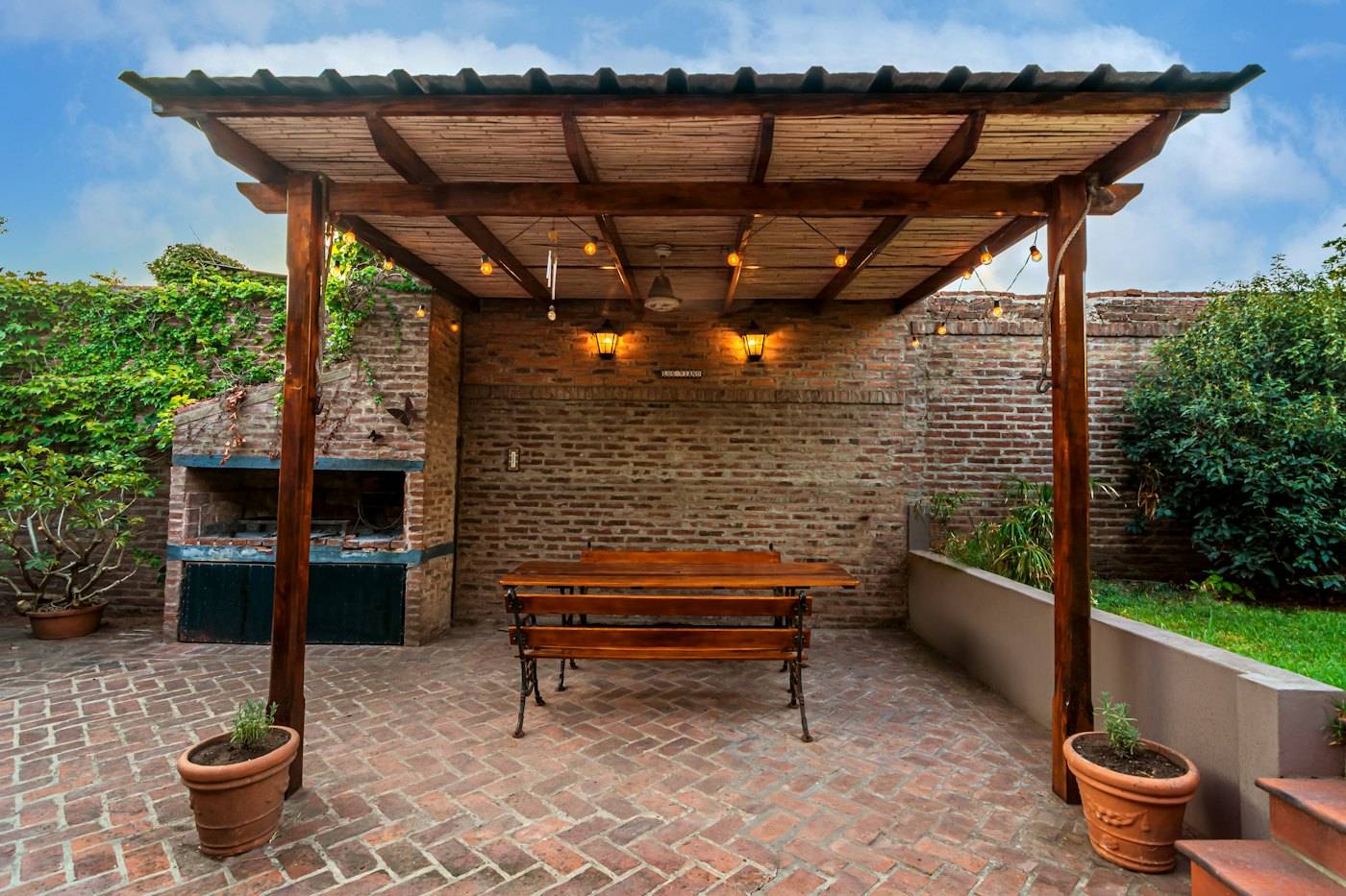 A timber pergola over a brick patio with a bench, a stone grill and planters