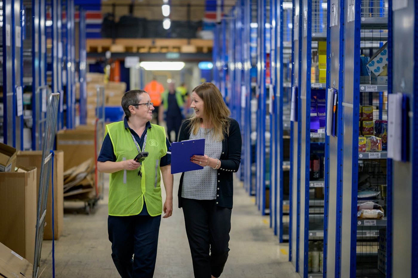 Two people talking as they walk down a warehouse aisle lined with blue shelving