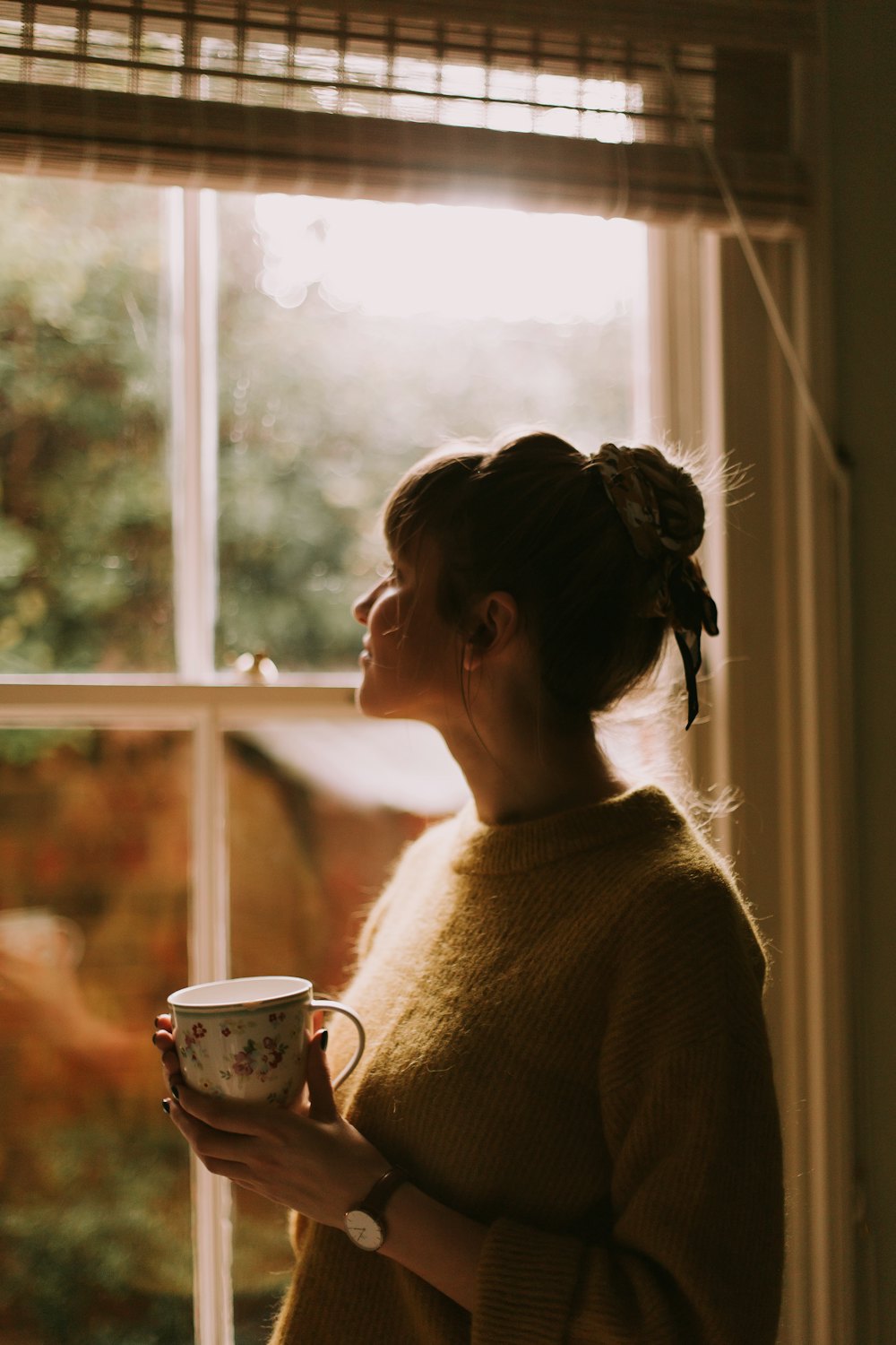A woman in a soft sweater holding a teacup by a sunny window