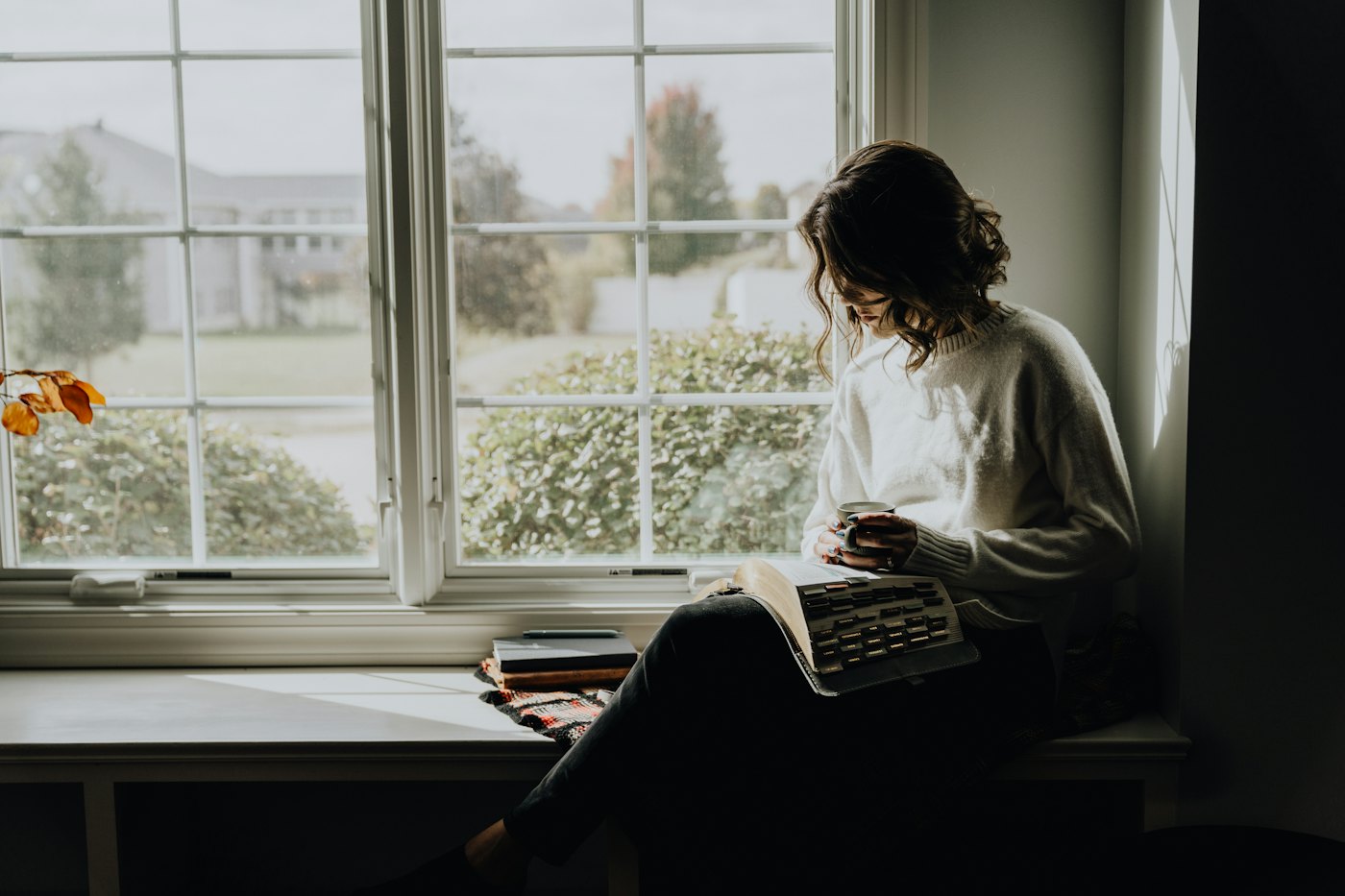 A woman with a warm drink reading quietly on a window seat